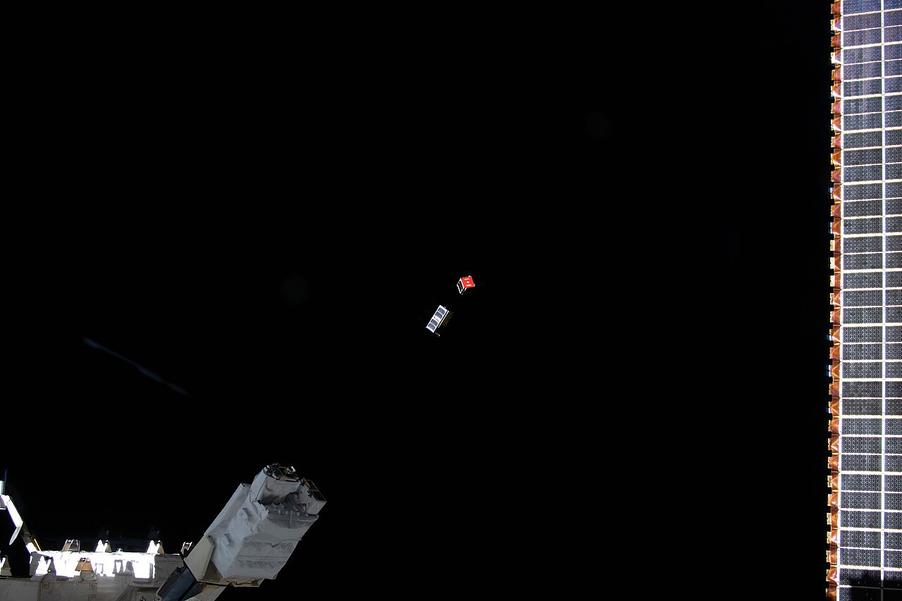 Two CubeSats floating beside the International Space Station against the blackness of space.